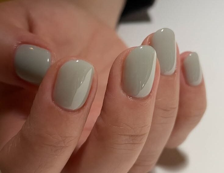 Mano con uñas cortas cuadradas con esquinas redondeadas, esmaltadas en un tono gris verdoso suave o verde menta apagado.
